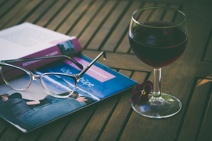 wine glass filled with liquor beside book and eyeglasses