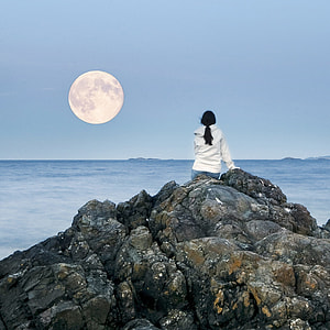 woman wearing white hoodie on top of rock facing towards the full moon