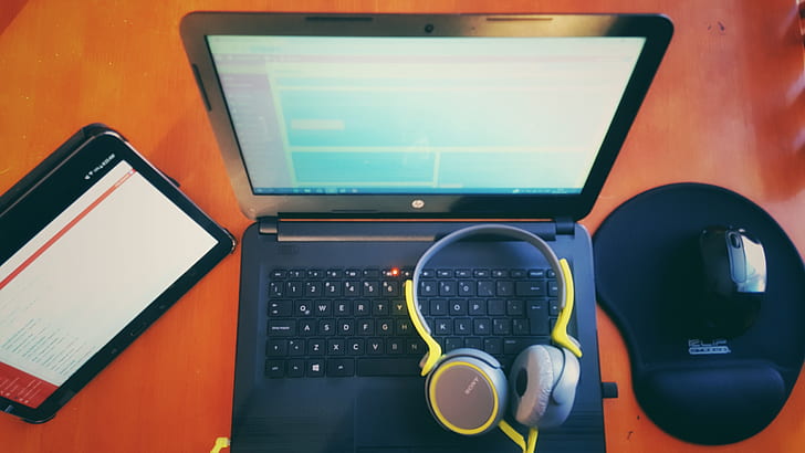 black laptop computer, wireless mouse, tablet computer, and yellow headphones on top of brown wooden furniture