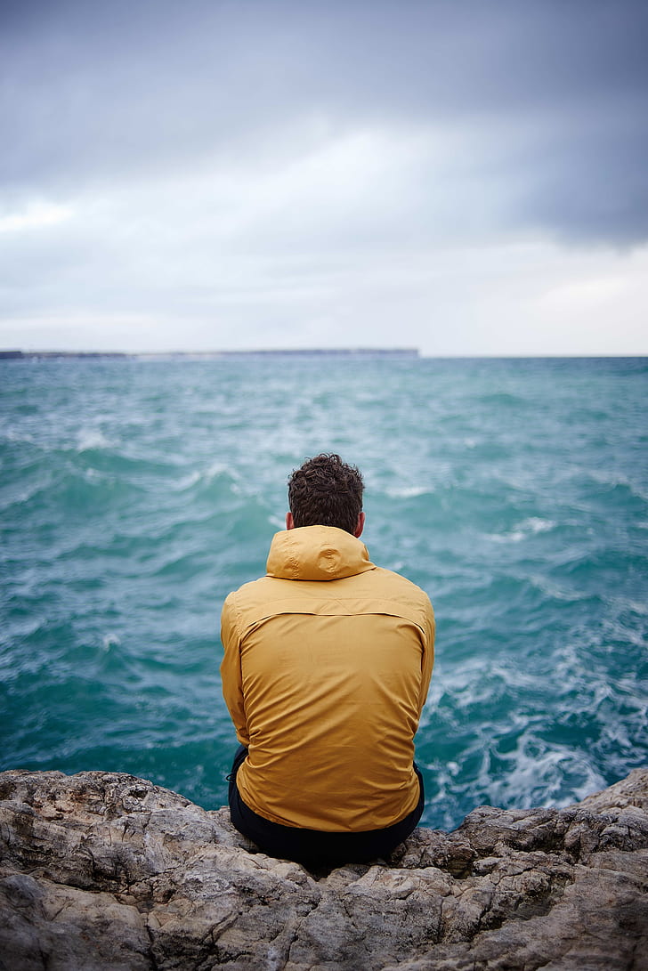 man in yellow hooded jacket sitting on cliff in front of sea