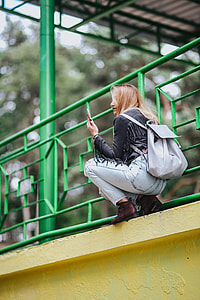Blonde woman in a black jacket and ripped jeans by a green handrail