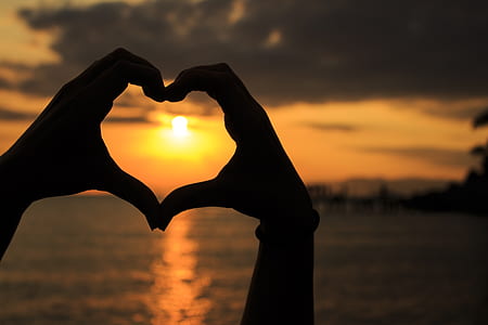 silhouette photo of person forming his two hands together a heart in seashore during sunset