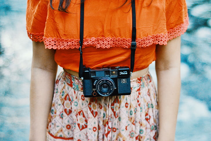 woman in orange top and white floral bottoms with black compact film camera