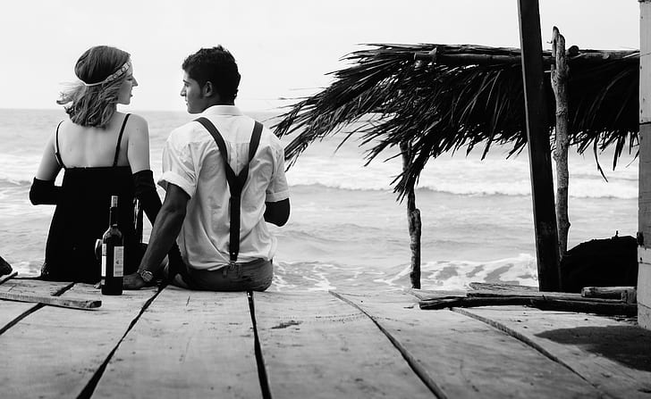 grayscale photo of couple sitting on wooden surface near seashore at daytime