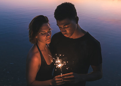 man and woman in black top holding together sparkler light near body of wtaer