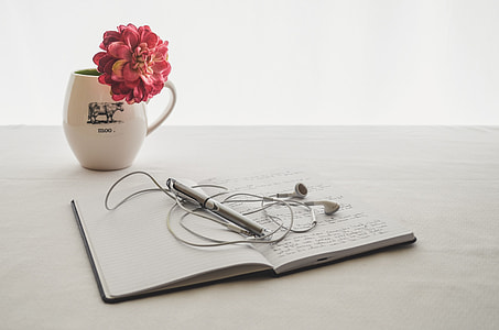 photo of opened notebook with pen and earphones with white ceramic mug and pink petaled flower in bloom