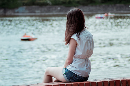 woman wearing white sleeveless dress sitting on brown brick near body of water during daytime