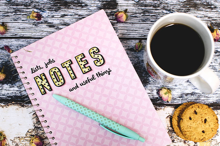 pink Notes with teal click pen next to white ceramic mug with coffee