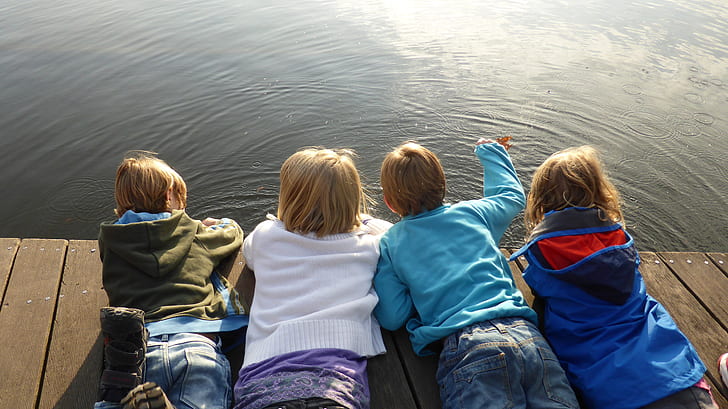 four children lying on brown deck while staring at body of water