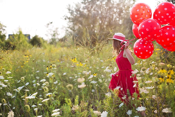 woman in red sleeveless dress holding red balloons walking on green plants during daytime
