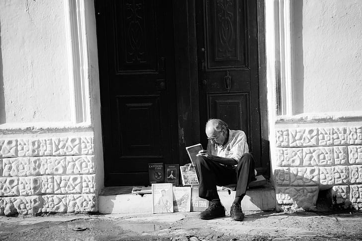 grayscale photo of man sitting on entrance displaying books