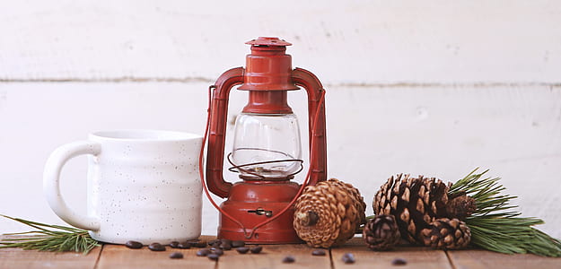 red kerosene lantern, white ceramic mug, and brown pine cones