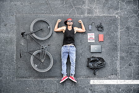 man wearing black tank top and blue denim jeans lying on road beside bike and laptop