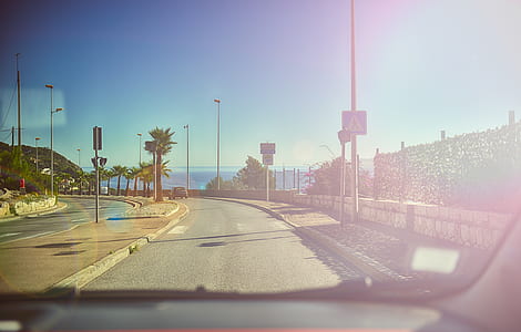 This picture is taken from inside a car through the windhield with the dashboard visible on the lower edge of the picture. The car is driving down a palm-lined ocean road under a blue sky with the ocean inthe background. The sunshine is reflected in the windshield of the car.