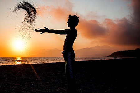 silhouette photography of man throwing sand near body of water during golden hour