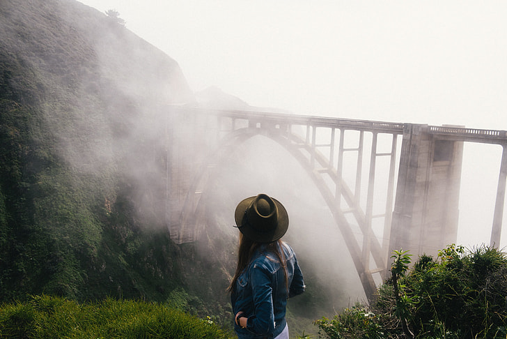 woman in cowboy hat and blue denim jacket standing on cliff with a view of concrete train bridge covered with fogs