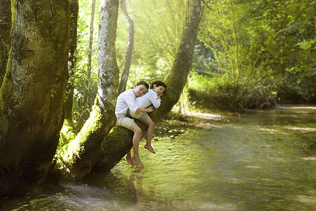 two boys sitting on tree branch above stream during day