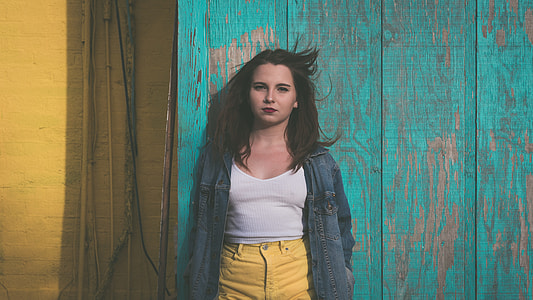 shallow focus photography of woman in white tank top and yellow jeans wearing blue denim jacket leaning on green wooden wall during daytime