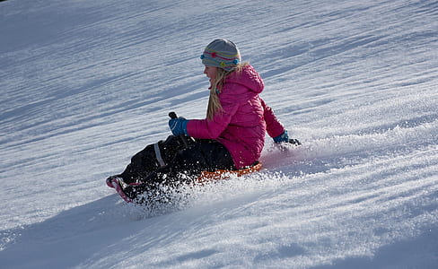 woman wearing pink zip-up hooded bubble jacket sits on orange sled on top of snow