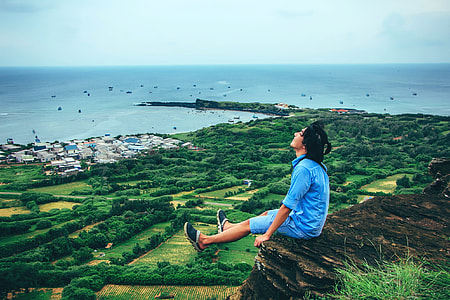 man sitting on cliff under blue clear skies during daytime at panoramic view photography