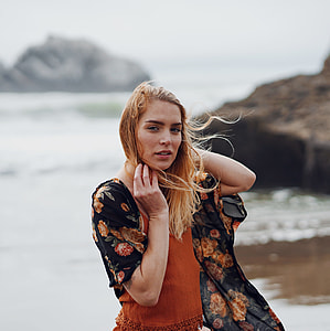 woman wearing orange spaghetti strap top with black and red floral cardigan standing on seashore with rocks during daytime