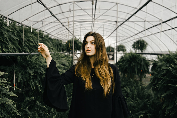 woman wearing black long sleeved dress inside greenhouse surrounded by green leaf plants