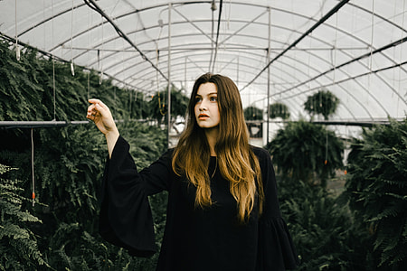 woman wearing black long sleeved dress inside greenhouse surrounded by green leaf plants