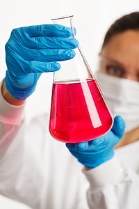 person in white laboratory gown and blue gloves holding clear glass laboratory flask with red liquid