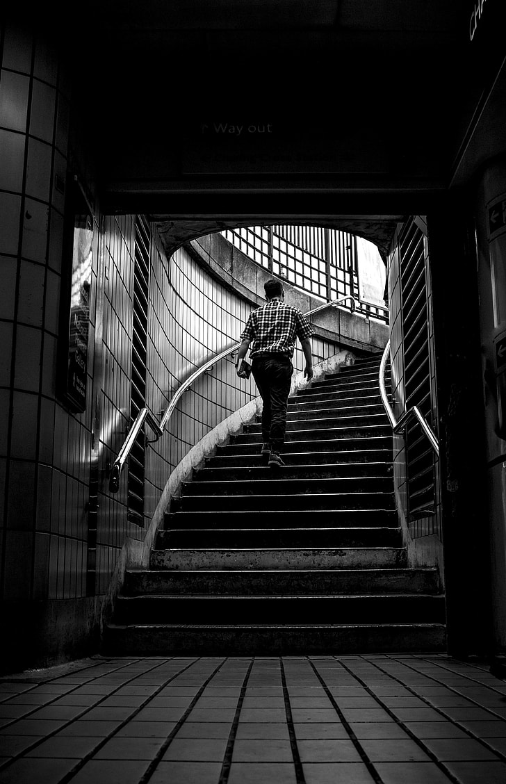 grayscale photo of man carrying books while walking on stairs at a subway station
