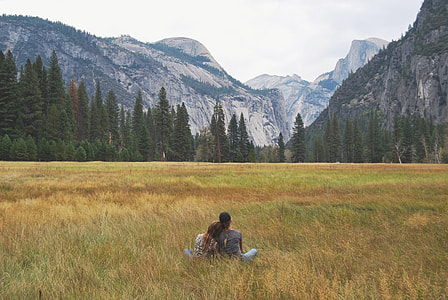 two person sitting on grass field facing alps across the forest during daytime