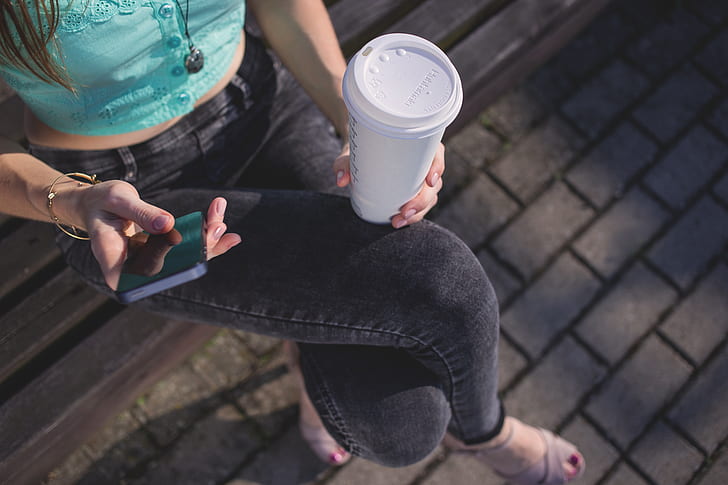 woman in black denim jeans holding white plastic tumbler sitting on brown wooden bench