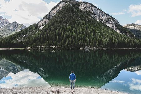 man standing in front of body of water near tree covered mountain during daytime