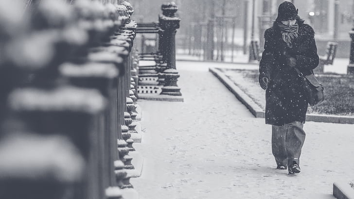 grayscale photography of a person in black trench coat walking across the street during winter weather