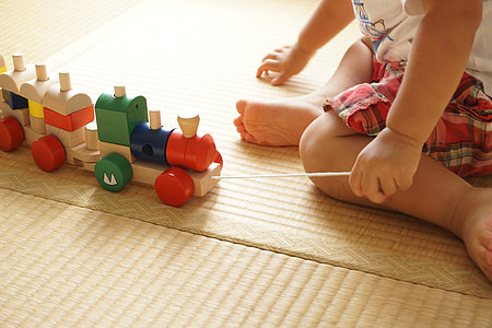 baby wearing white shirt holding assorted-color wooden train toy sitting on brown wooden flooring