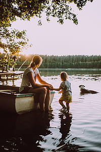 woman wearing white t-shirt and black denim shorts sitting on boat while holding kid wearing white t-shirt on lake with brown dog at distance during daytime