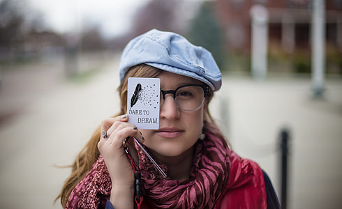 woman wearing pink and maroon zebra pattern scarf, red and black shirt, black and white plastic framed eyeglasses and blue hat while holding white Dare To Dream printed paper during daylight
