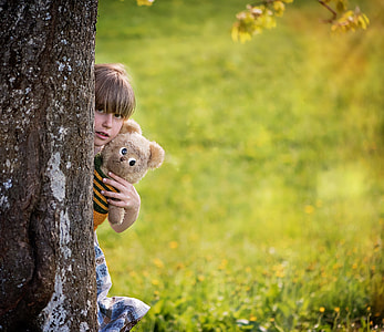 child hiding behind the tree while holding brown bear plusht oy