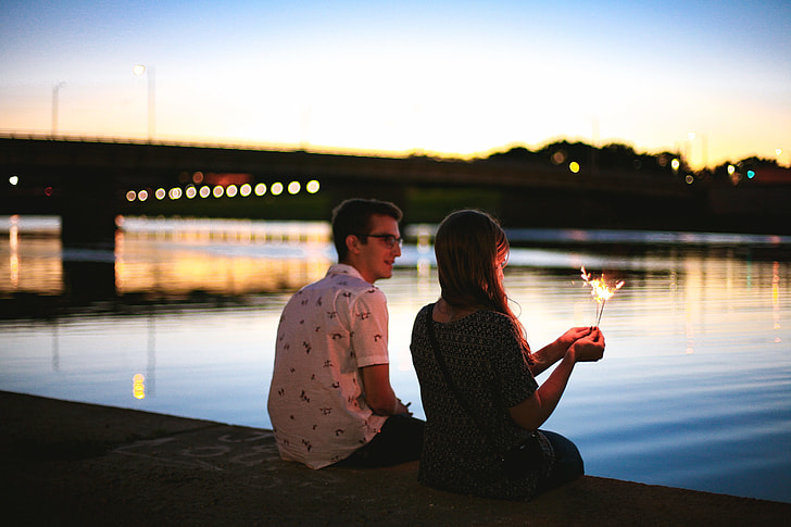 man and woman sitting beside river near bridge during sunset