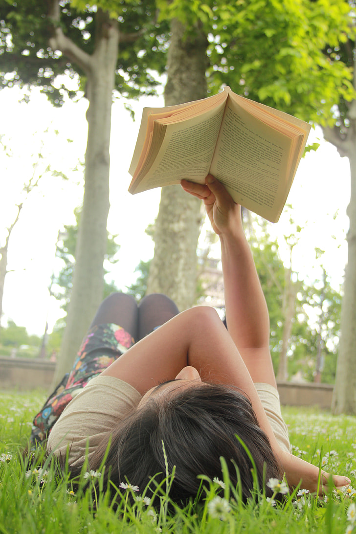 woman lying on grass field reading book beside green tree