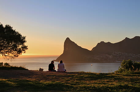 man and woman sitting on green grass field watching mountain and ocean view during golden hour