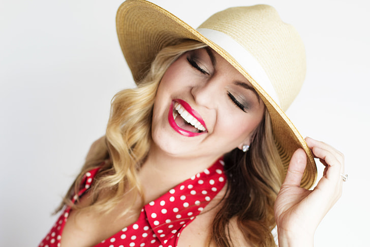 woman in red and white polka-dot blouse smiling white holding brown straw sun hat