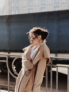 woman wearing brown coat leaning against metal fence during daytime