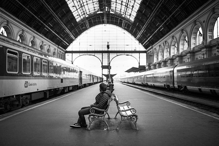 grayscale photo of man sitting on bench on train station