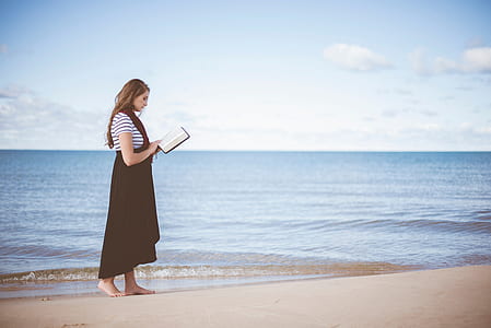 woman reading a book while standing on beach sand during daytime