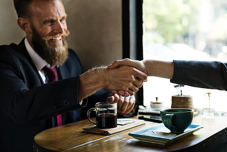 man in black suit in front of coffee on table shaking hands on person in other side