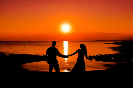 silhouette of man and woman holding hand stands near ocean at sunset