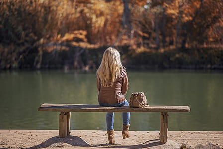woman wearing brown leather jacket and blue jeans sitting on bench