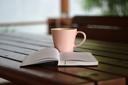 close-up photo of white labeled book open beside pink ceramic coffee mug on brown wooden table