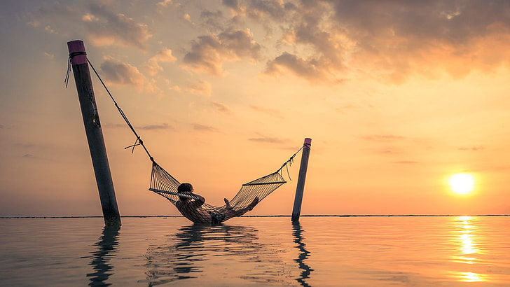 man lying in hammock above body of water during sunset