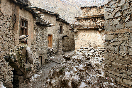 male soldier in gear set standing in front of rocky buildings
